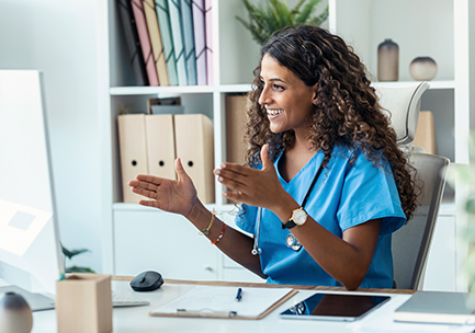 A smiling healthcare professional in blue scrubs sits at a desk, gesturing with her hands during a video call. A clipboard, smartphone, and plants are on the desk, and shelves with files are in the background.