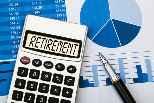 A calculator displaying the word RETIREMENT sits on top of financial documents with charts, graphs, and a pen, suggesting financial planning for retirement.