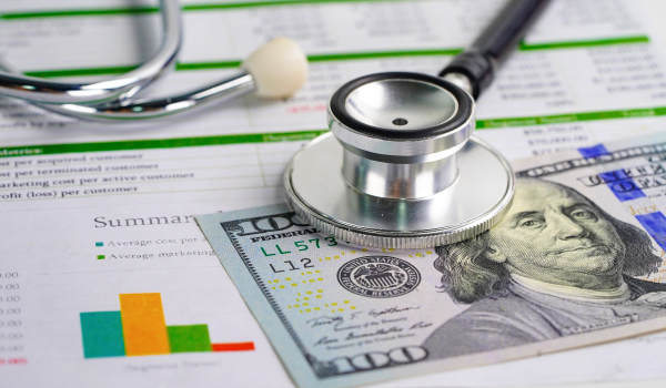 A stethoscope rests on a hundred-dollar bill placed over financial charts, symbolizing the relationship between healthcare and financial costs.