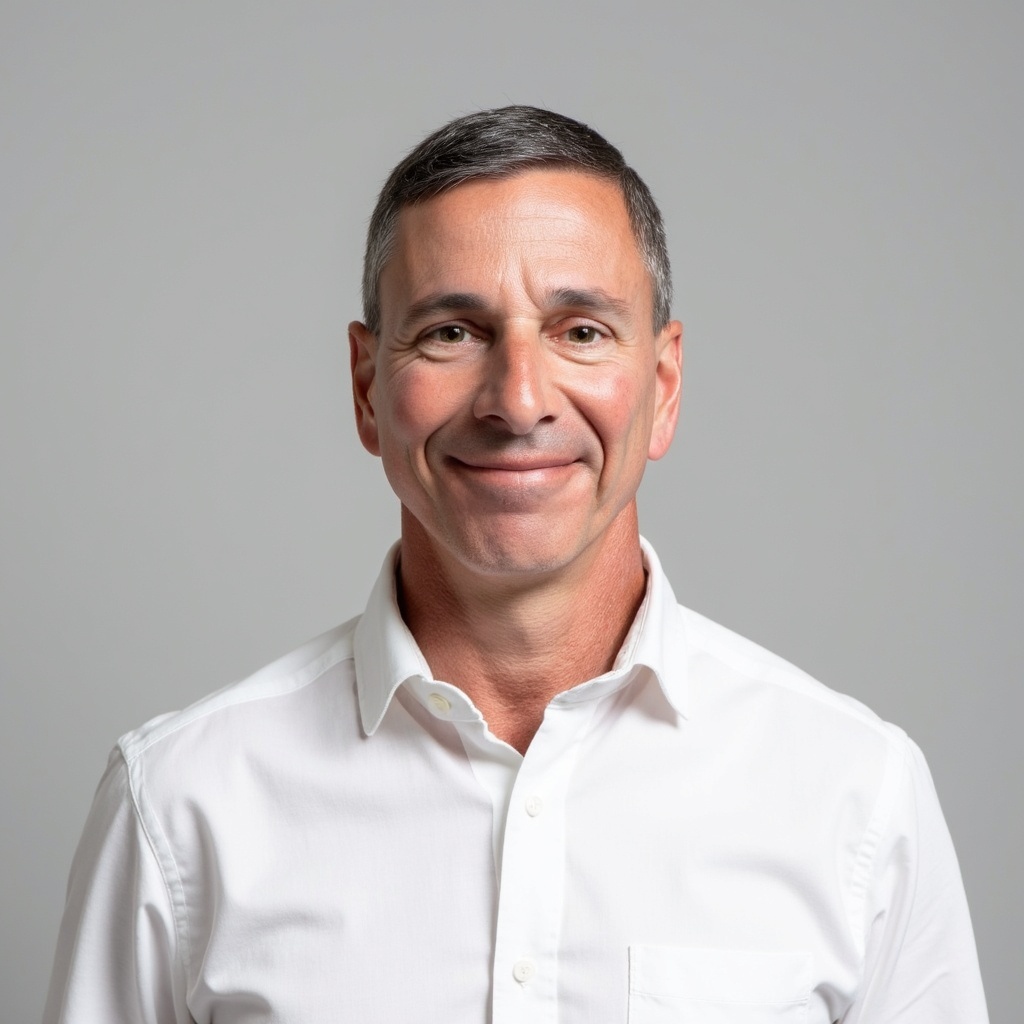 A middle-aged man with short dark hair, wearing a white button-up shirt, smiles at the camera against a plain light gray background.