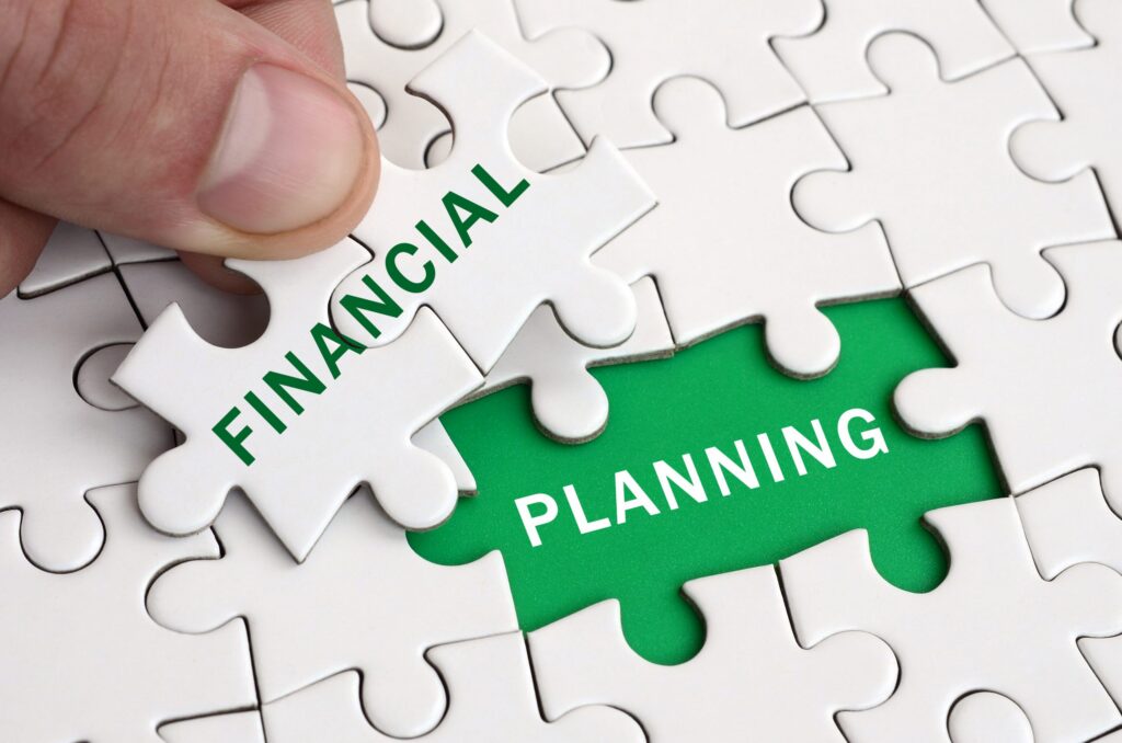 A close-up of a hand placing a puzzle piece labeled FINANCIAL to fit with another piece labeled PLANNING, symbolizing the concept of financial planning. The pieces complete a puzzle over a green background.