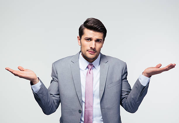 A man in a gray suit and pink tie stands against a light background, raising both hands with palms up and a confused or uncertain expression on his face.