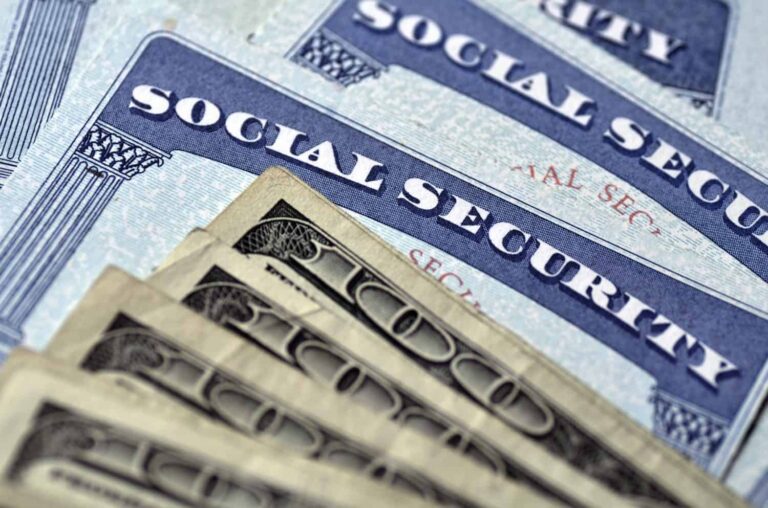 Several Social Security cards are layered above a spread of $100 bills, symbolizing the relationship between Social Security benefits and financial security or income.